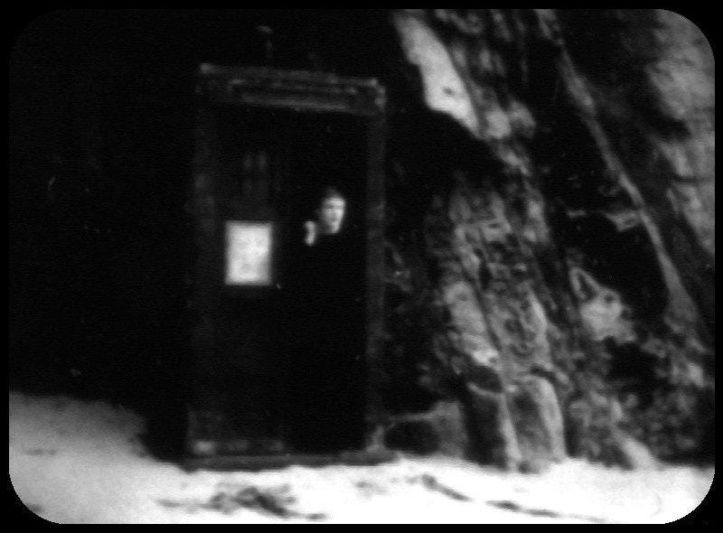 A monochrome photograph taken from a television screen showing a cave entrance from the outside with the TARDIS prop next to it with its door open and an indistinct white-skinned figure peering out. 
