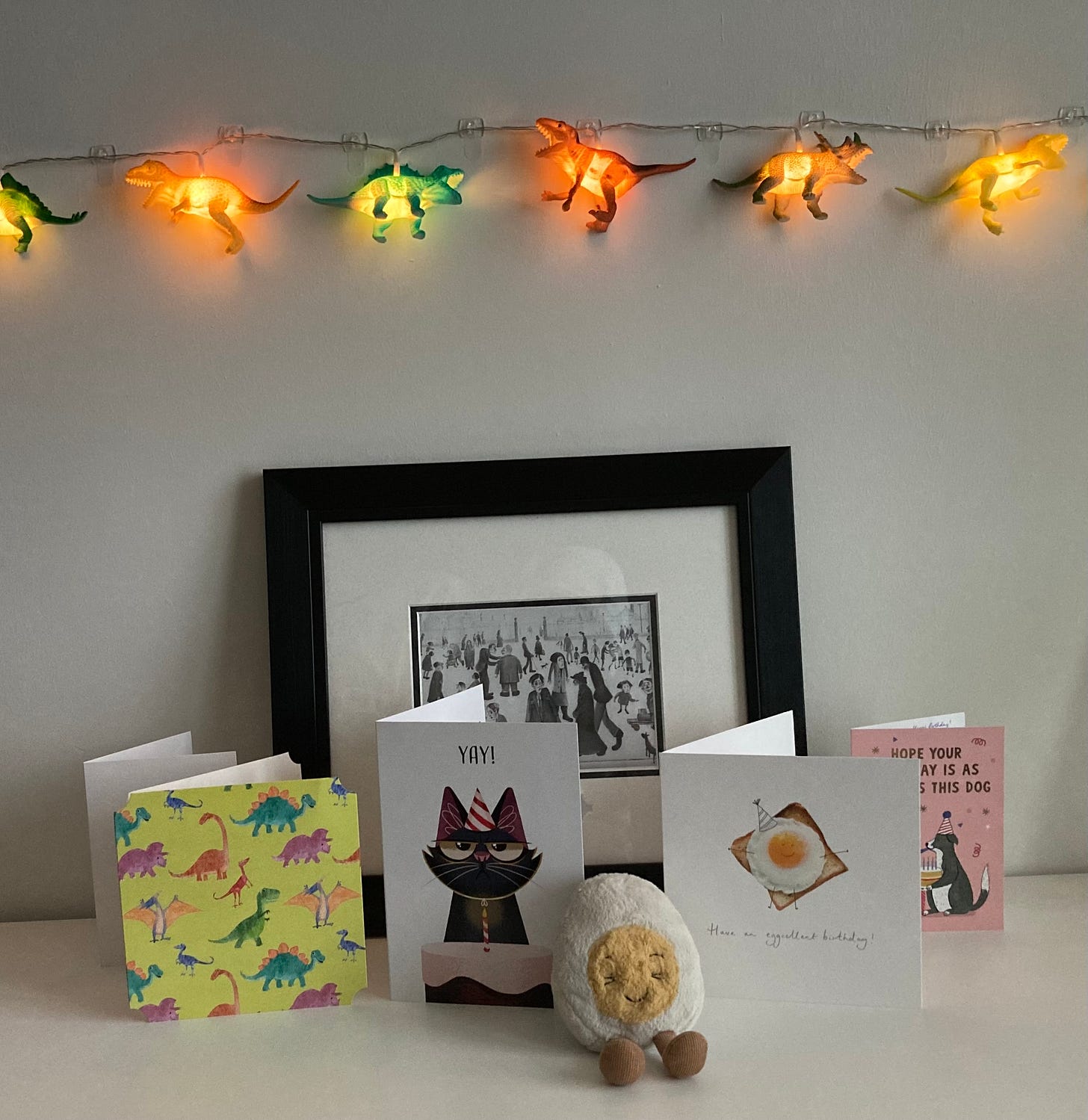 Dippy the soft toy egg sitting in front of four birthday cards, one of which features a fried egg, and a print of LS lowry's artwork 'The Cripples'. Dinosaur fairy lights are hanging on the wall, roaring at eachother. 