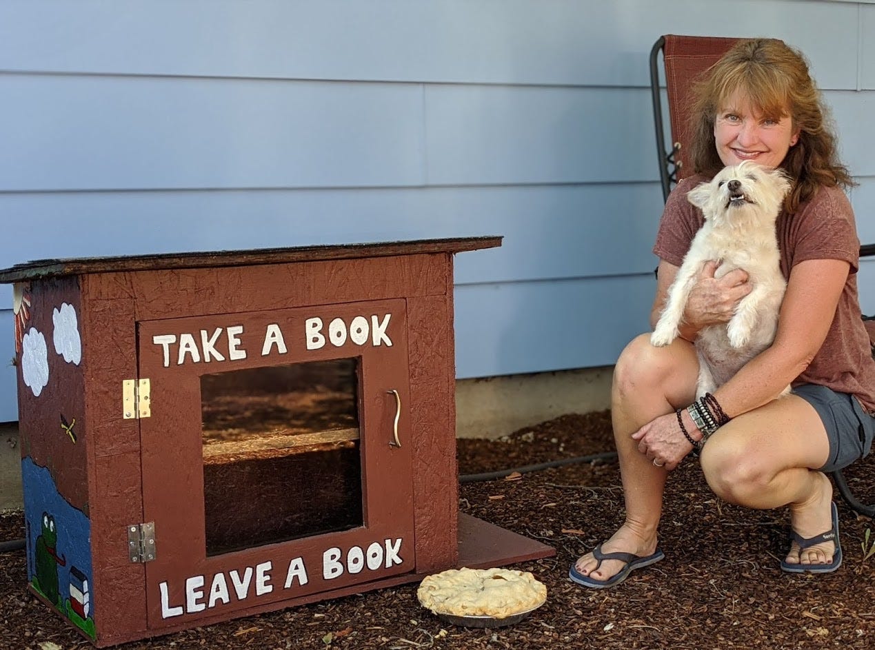 Brown haired woman in shorts holding small white dog next to red little free library and apple pie.