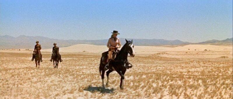 Carrie rides a horse in the desert as two men trail behind her