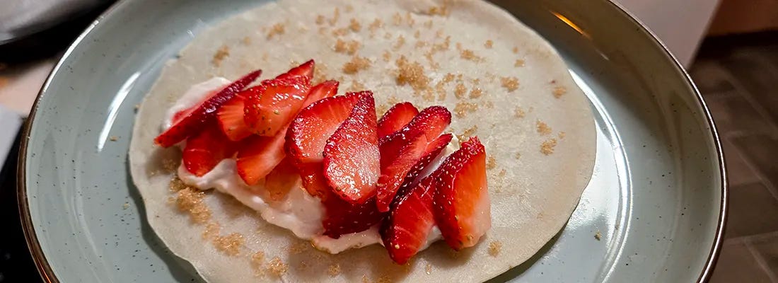 A thin crepe or tortilla on a plate topped with a layer of cream and neatly arranged slices of fresh strawberries, with a light sprinkle of brown sugar over the surface.