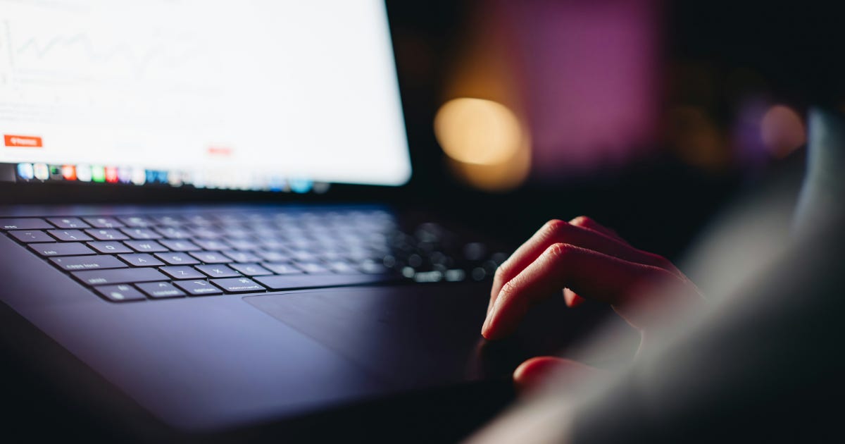 A person using a laptop in low light, with their hand resting on the trackpad.