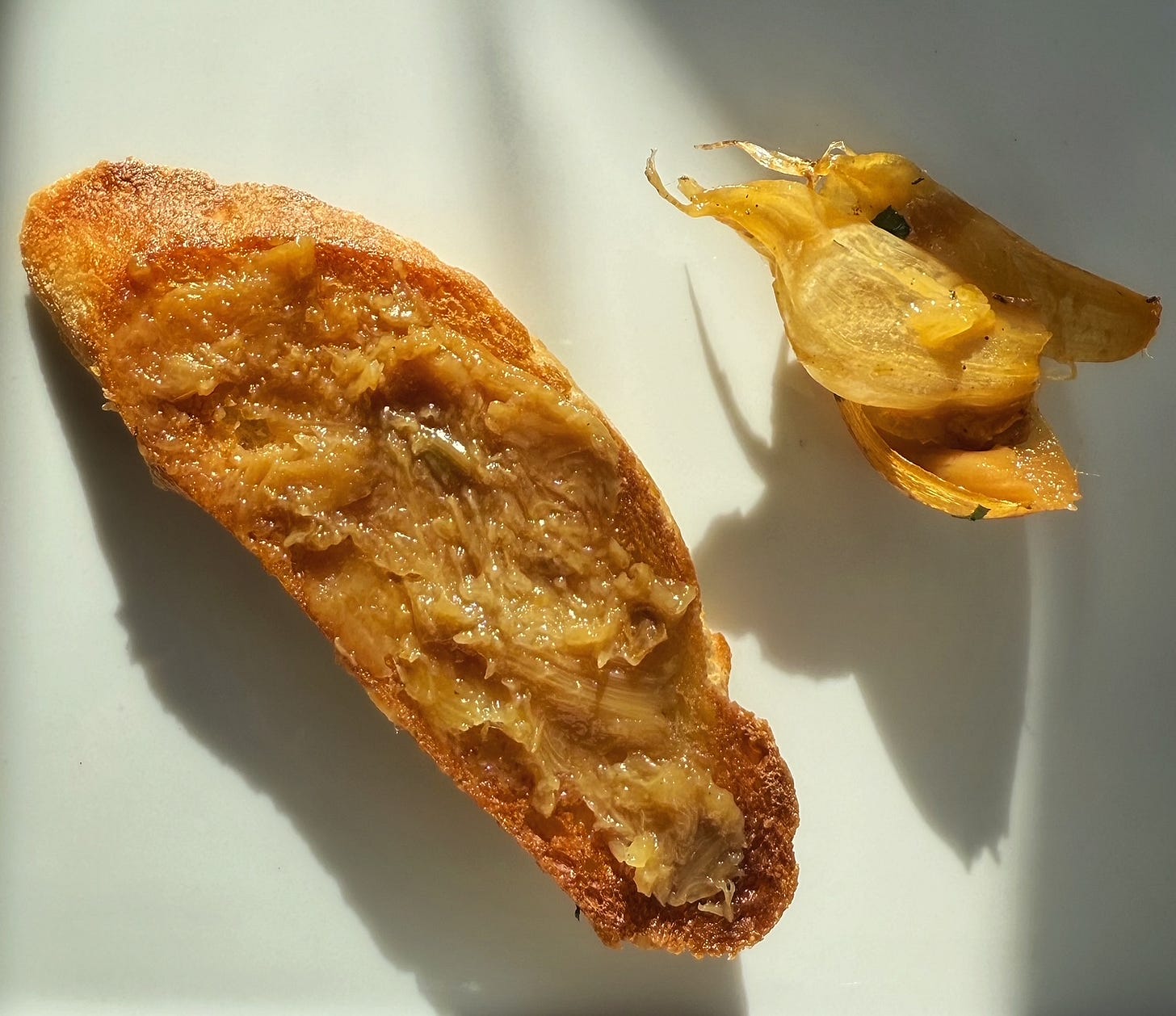 A toasted slice of baguette with sweet, roasted garlic slathered upon it. Spent garlic skin sits next to it. 