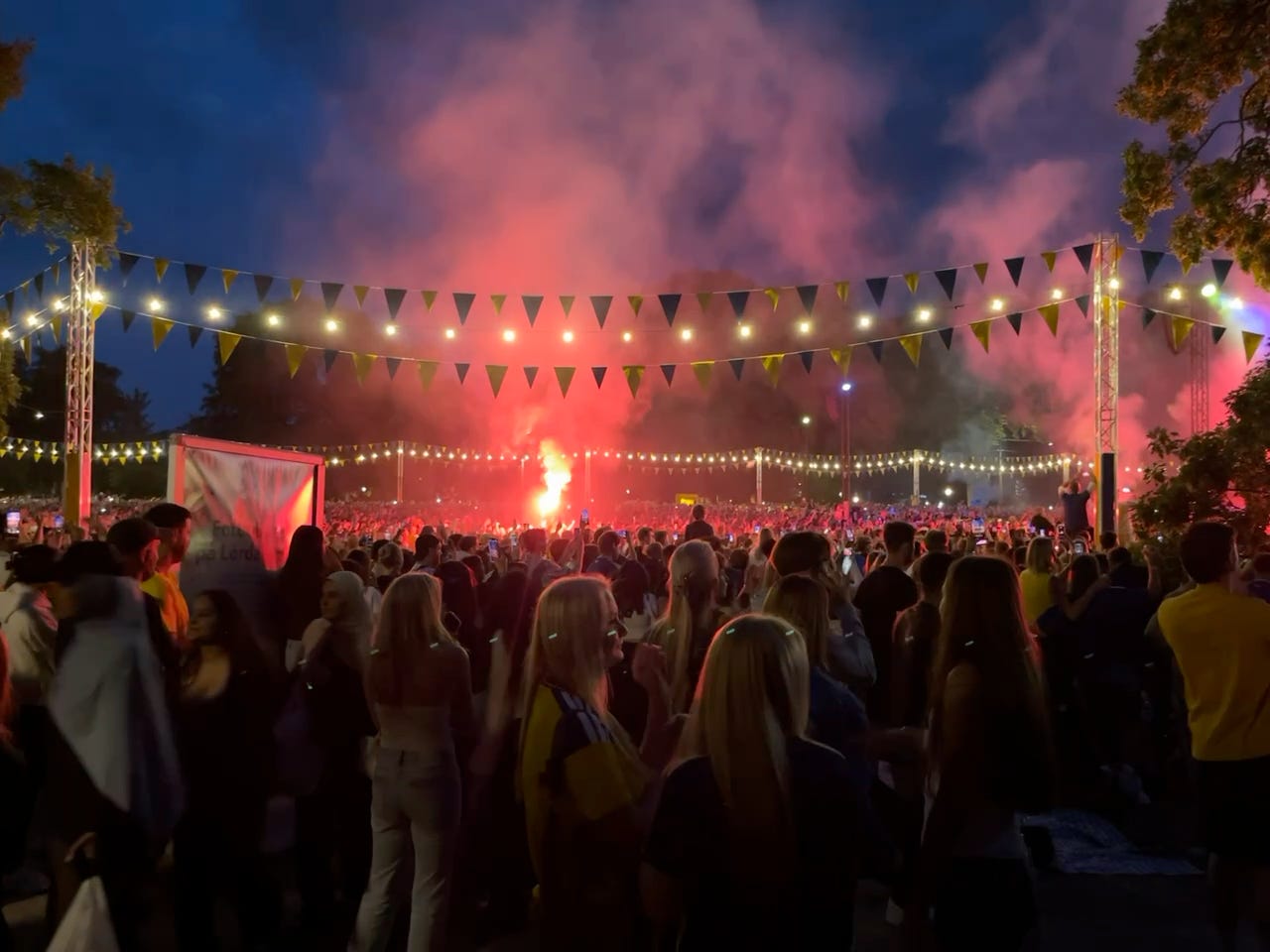 A dense crowd some wearing the Swedish national soccer team's yellow jersey. There are flares in the distance, with smoke covering large portions of the dark blue night sky. There are lines of yellow and blue triangle flags hanging over the crowd, as well as lines of round lights