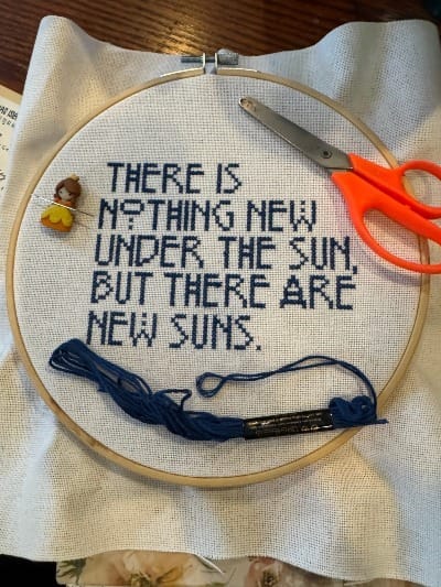 A cross stitch project that's partially complete. Dark blue words on a white fabric say There is nothing new under the sun but there are new suns