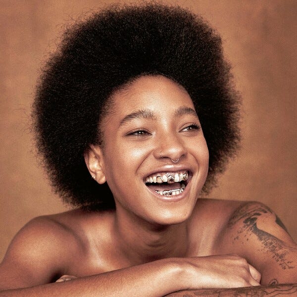 Willow Smith poses with an Afro. In one photo, she is wearing a plaid jacket during a red carpet appearance. In the second photo, she smiles widely with grills on her teeth for the empathogen album cover.