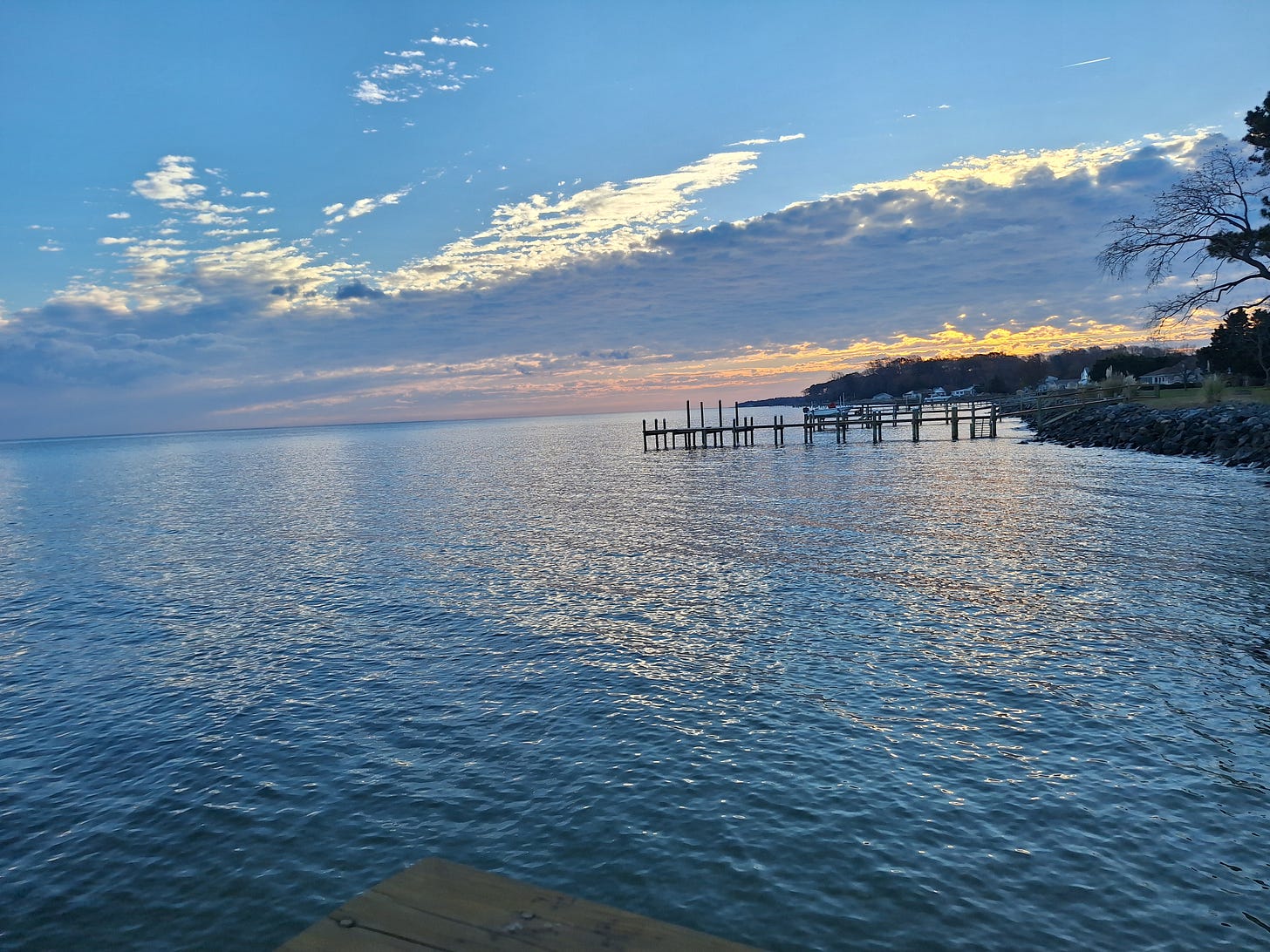Still water under a soft morning sky with gentle clouds and calm light — a peaceful, quiet scene with a wooden pier and shoreline in the distance.