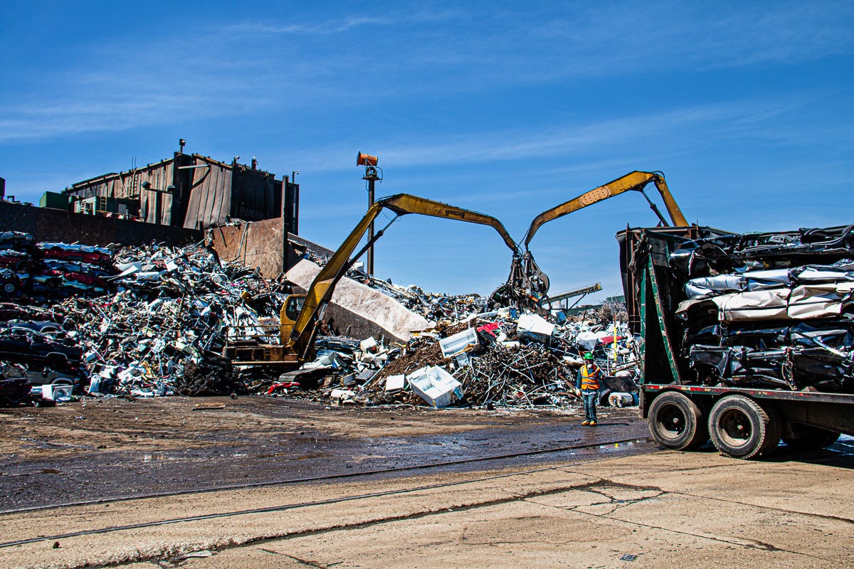 Two cranes actively digging into a massive heap of crushed and twisted metal in a large scrap yard under blue sky.