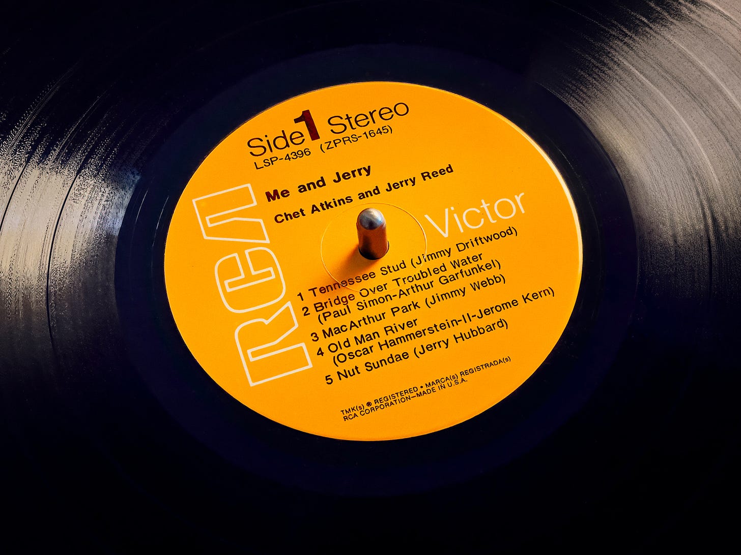 Close-up of the orange RCA Victor Side 1 label on the Me and Jerry LP by Chet Atkins and Jerry Reed, the record tilted on a dark surface with light raking across the grooves. The listing reads Tennessee Stud, Bridge Over Troubled Water, MacArthur Park, Old Man River, and Nut Sundae — the last credited to Jerry Hubbard, Reed's given name. Color photograph by Sean Perry for The Sunday Find.