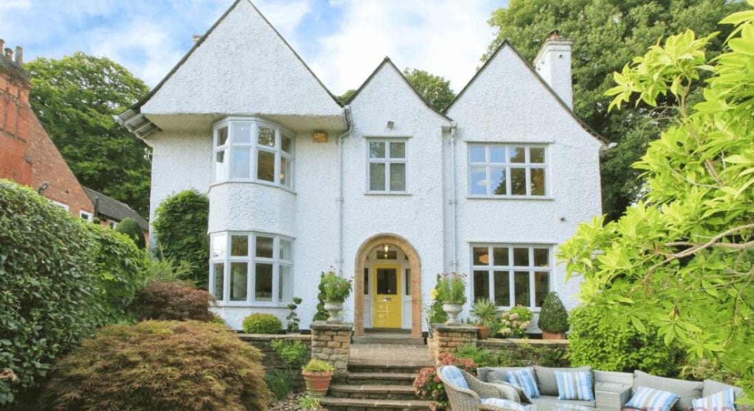 A white house with a yellow door, grey stone steps, and hedges either side of it