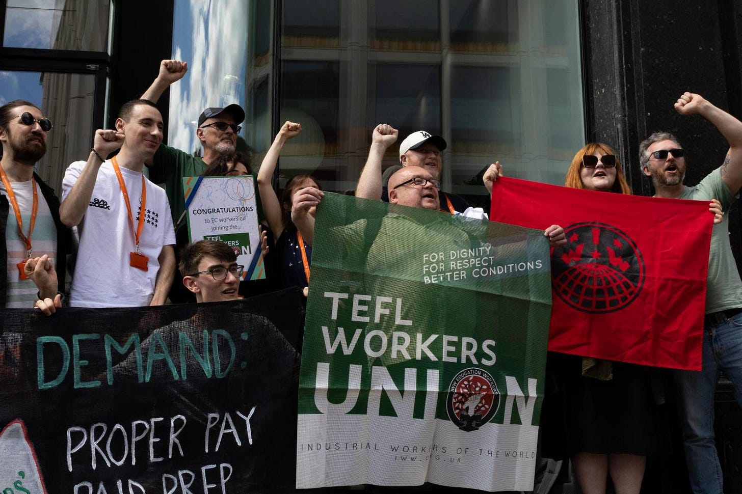 Photograph of a group of TEFL workers rallying outside EC English London location, demonstrating their readiness for strike action by raising their fists in the air while hodling signs and IWW banners. Photograph of a group of TEFL workers rallying outside EC English London location, demonstrating their readiness for strike action by raising their fists in the air while hodling signs and IWW banners.
