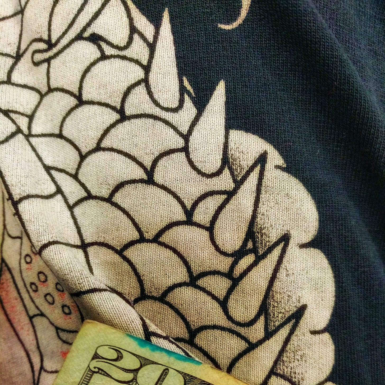 close-up of cotton fabric with various patterns outlined in black on off-white of scales and teardrop shaped horns and a dark navy background on the right side; also the corner of a 20 dollar bill can be seen at the bottom edge of the image.