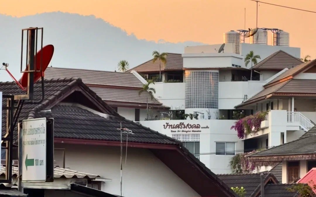 Tile-roofed Thai houses in the foreground with a white multi-storey condominium building behind, the Doi Suthep mountains and a golden sunset sky above, in a residential part of Chiang Mai.
