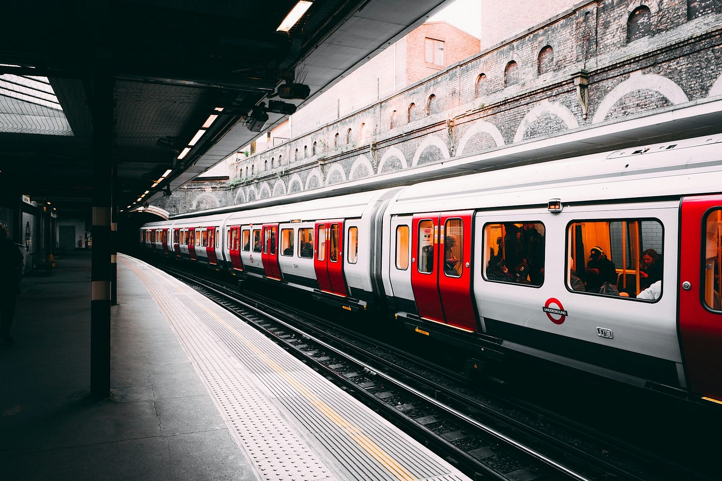 A London Underground train with red and white carriages stopped at a station platform, with passengers visible through the windows and classic brick architecture featuring arched recesses in the background.