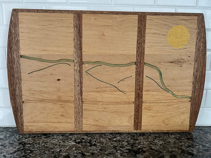 A series of mixed-media cards hang on a beaded string and in a frame, a painting of a shipwreck ocean scene with a wooden frame, an abstract view through my dining room window of the mountain carved into a wooden cutting board, and a patchwork quilt laying atop a couch.
