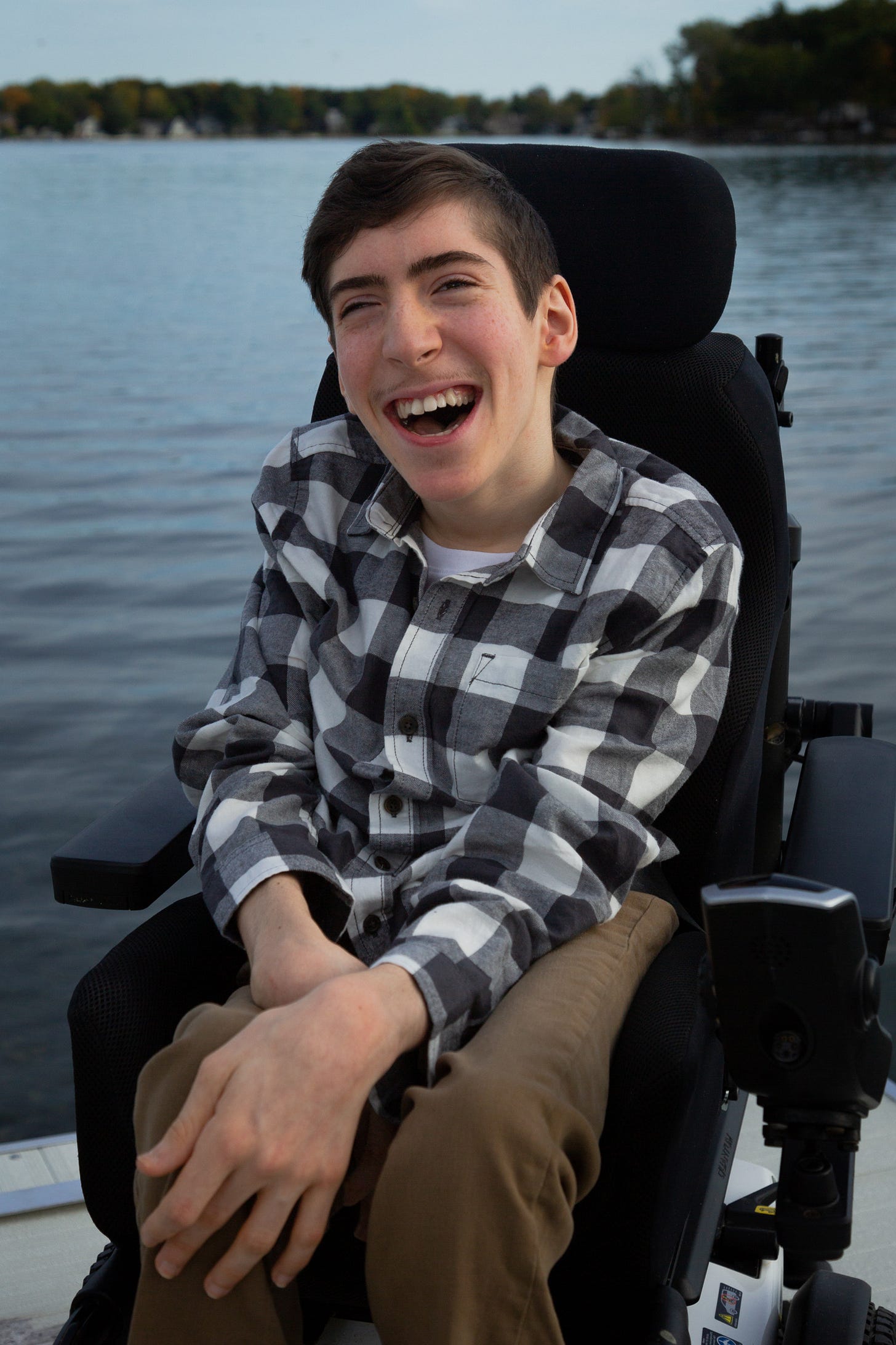 Enzo Cascardo smiles broadly while sitting in his black power wheelchair on a boat. He wears a black-and-white plaid shirt and brown pants. Calm water and a tree-lined shoreline create a peaceful backdrop. Enzo Cascardo smiles broadly while sitting in his black power wheelchair on a boat. He wears a black-and-white plaid shirt and brown pants. Calm water and a tree-lined shoreline create a peaceful backdrop.