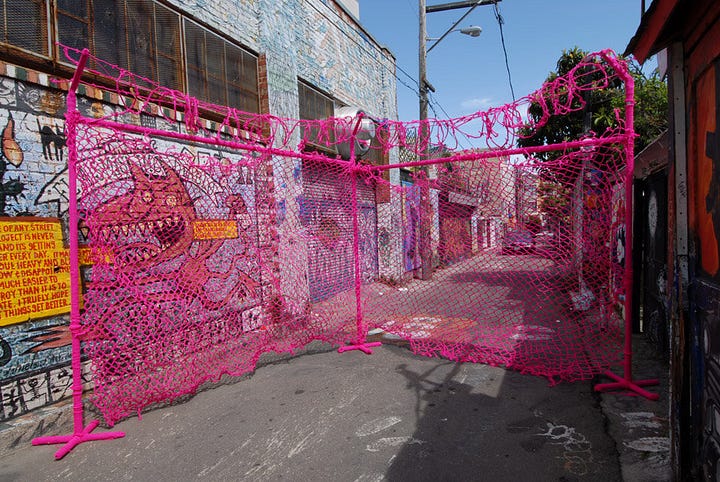1) A large knit pink chain-link fence with knit barbed wire is seen with people outside of it, looking in. 2) The fence is now outside, in an alleyway, with a graffiti-ed wall to the left.