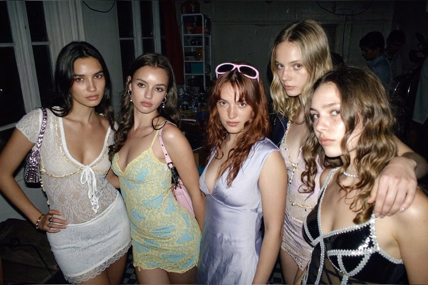 A group of young women friends, mixed ethnicities, wearing short party dresses, smiling and laughing with their arms around each other at a house party in Shoreditch, London.