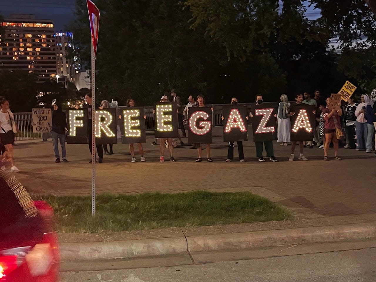 A group of people hold up signs that spell out FREE GAZA in twinkle lights at dusk on a city street corner.
