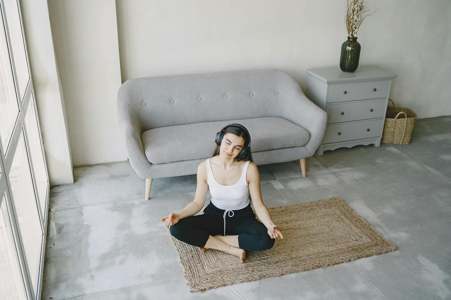 Person sitting cross-legged on a woven mat, wearing headphones and practicing meditation in a calm, minimal living space.