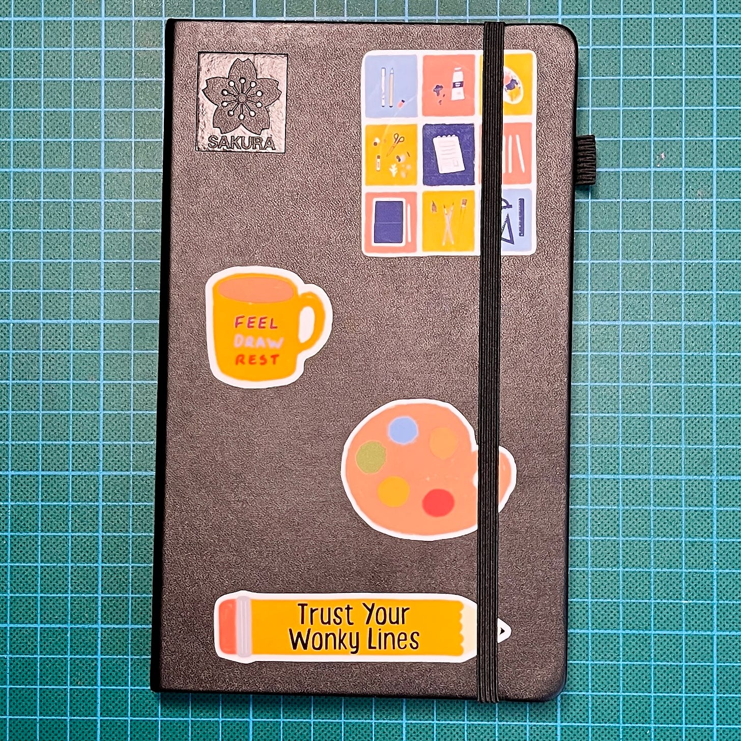 Photo d'un sketchbook posé sur mon tapis de découpe : la couverture est noir, on voit le logo Sakura ainsi que plusieurs autocollants. Photo d'un sketchbook posé sur mon tapis de découpe : la couverture est noir, on voit le logo Sakura ainsi que plusieurs autocollants.