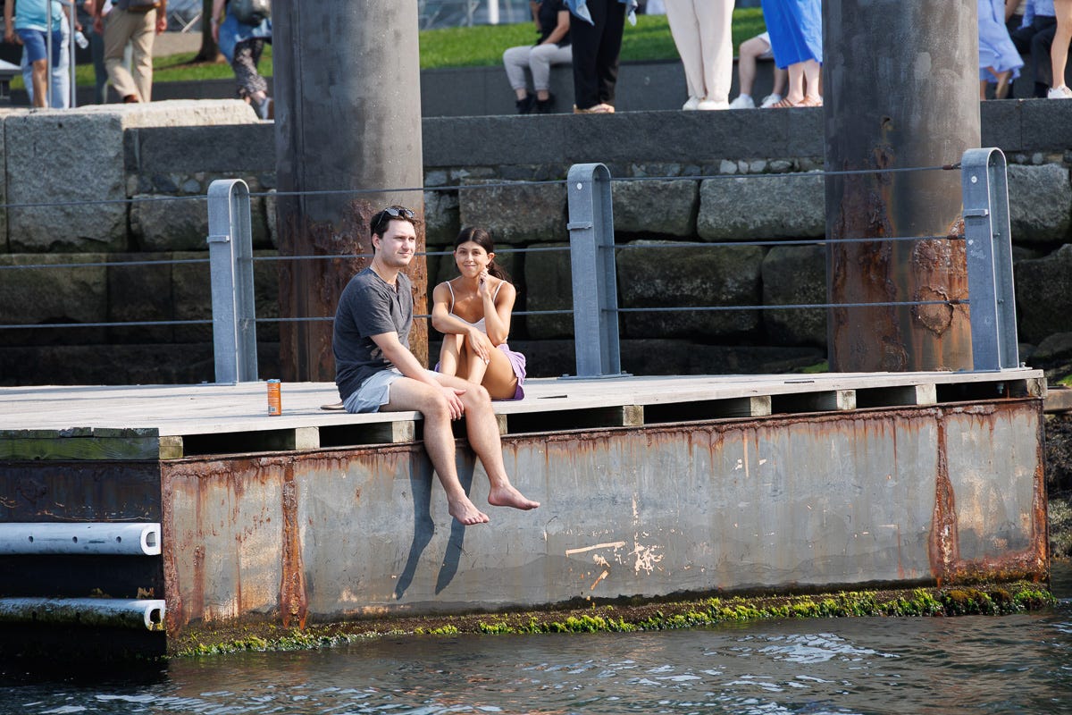 Two people sitting on dock edge in Boston Harbor, talking with feet hanging above the water