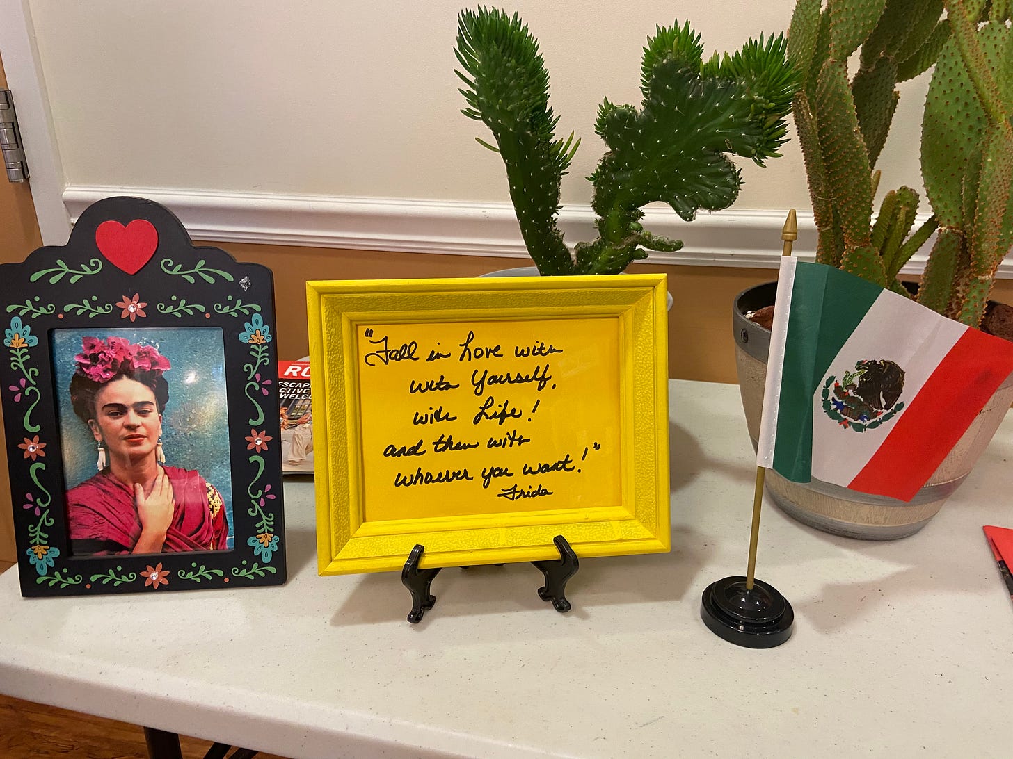 a photo of a cactus, Mexican flag, picture of Frida Kahlo, and a quote by her: "Fall in love with yourself, with life and then with whoever you want!"