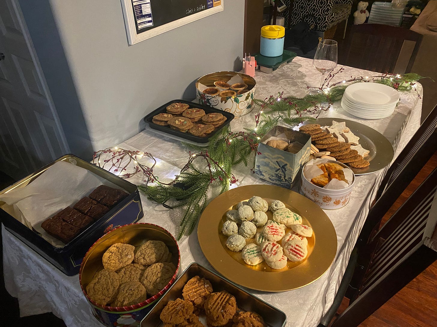 A table with a white tablecloth, a fake evergreen garland, and a string of small lights, showcasing plates and containers of at least a dozen different types of cookies and bars.