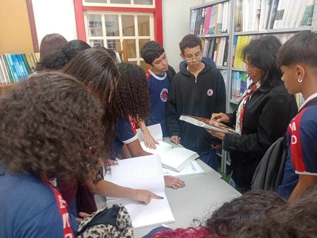#descrevemospravocê  Fotografia feita dentro de uma biblioteca mostra um grupo de estudantes reunido ao redor de uma mesa, observando livros com atenção. Os jovens vestem uniforme escolar azul-marinho com detalhes vermelhos e brancos. No centro da imagem, uma mulher adulta, de cabelos escuros e lisos, veste um casaco preto e segura um livro aberto, explicando algo para o grupo. Ela parece conduzir a atividade, provavelmente uma mediação de leitura ou apresentação de acervo.  Os estudantes demonstram interesse: alguns folheiam os livros sobre a mesa, enquanto outros olham para a mulher que fala. Ao fundo, prateleiras metálicas organizadas com livros e documentos encadernados. À esquerda, uma parede clara com livros coloridos e uma porta de moldura vermelha completam o cenário. A cena transmite um momento de aprendizagem coletiva e aproximação com o universo dos livros em ambiente escolar ou cultural.