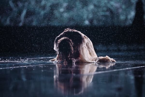 A photograph of Taylor Swift, kneeling with her head to the floor, during her Eras Tour. She is soaked by the pouring rain.