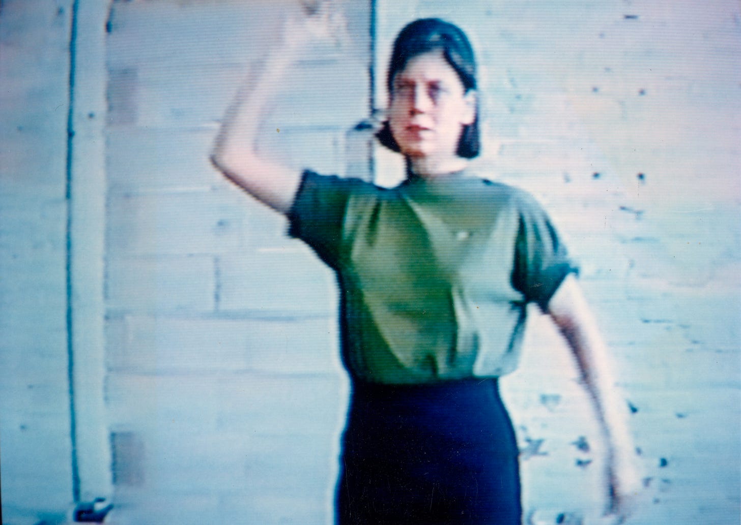 Lo-res video still of the artist, Mary Addison Hackett standing in an empty room facing the camera. Her right hand is raised overhead as she prepares to throw a dart. She’s wearing a short-sleeve green shirt and black pants. Lo-res video still of the artist, Mary Addison Hackett standing in an empty room facing the camera. Her right hand is raised overhead as she prepares to throw a dart. She’s wearing a short-sleeve green shirt and black pants.
