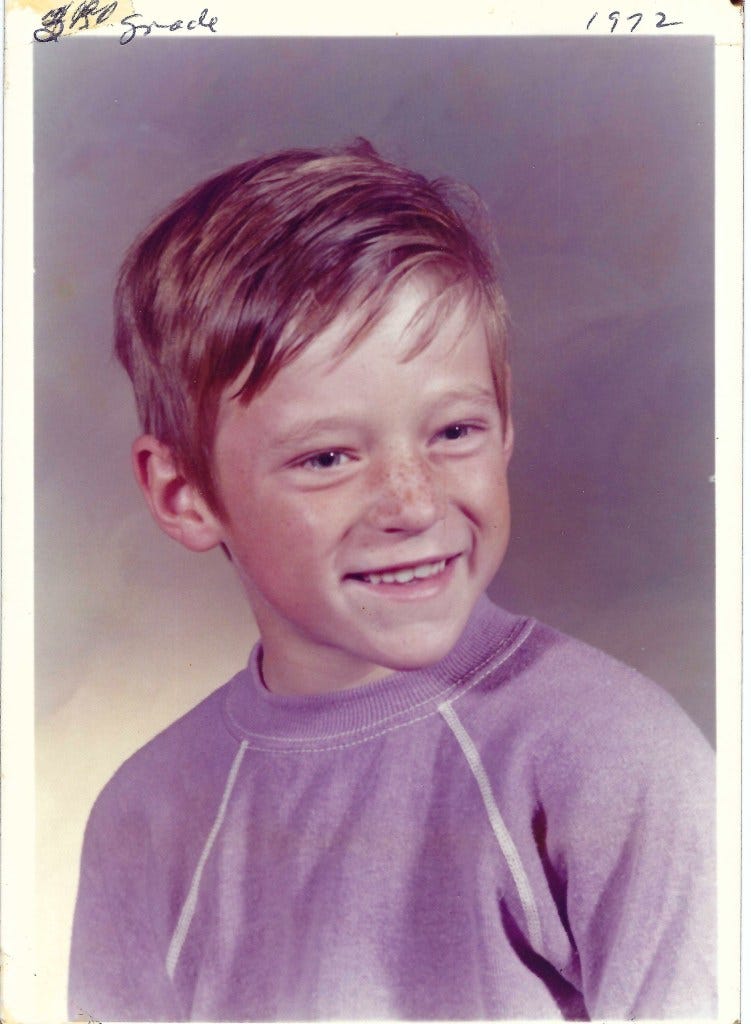 A young boy with short hair and freckles smiles for a school portrait, wearing a purple sweater. The photo is dated 1972.
