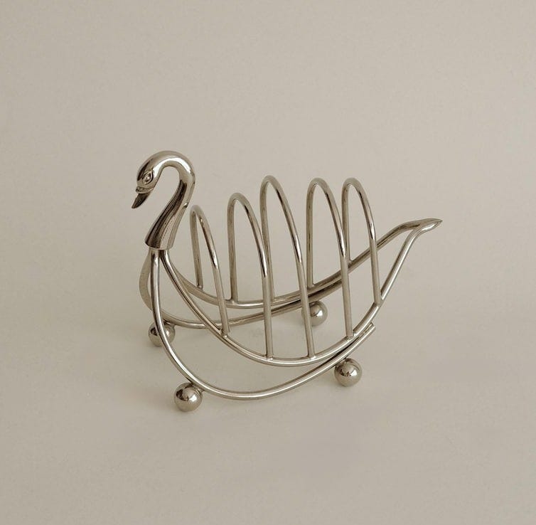 May include: A silver-toned metal toast rack shaped like a swan. The rack features a curved neck and head at the front, a series of vertical wire dividers for holding slices of toast, and four small spherical feet that provide a stable base.