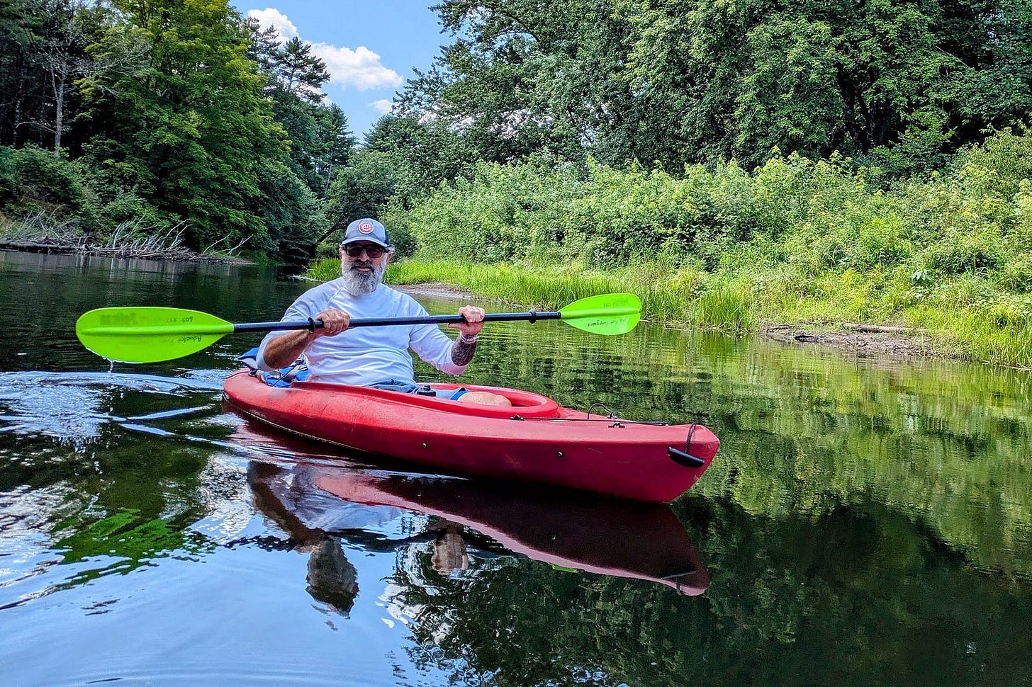 The author paddling a red kayak with bright green paddle blades on a calm river in New Hampshire. He wears a white long-sleeve shirt, gray cap, and sunglasses. Lush green trees and shrubs line the riverbank, their reflections mirrored in the still water under a summer sky. The author paddling a red kayak with bright green paddle blades on a calm river in New Hampshire. He wears a white long-sleeve shirt, gray cap, and sunglasses. Lush green trees and shrubs line the riverbank, their reflections mirrored in the still water under a summer sky.