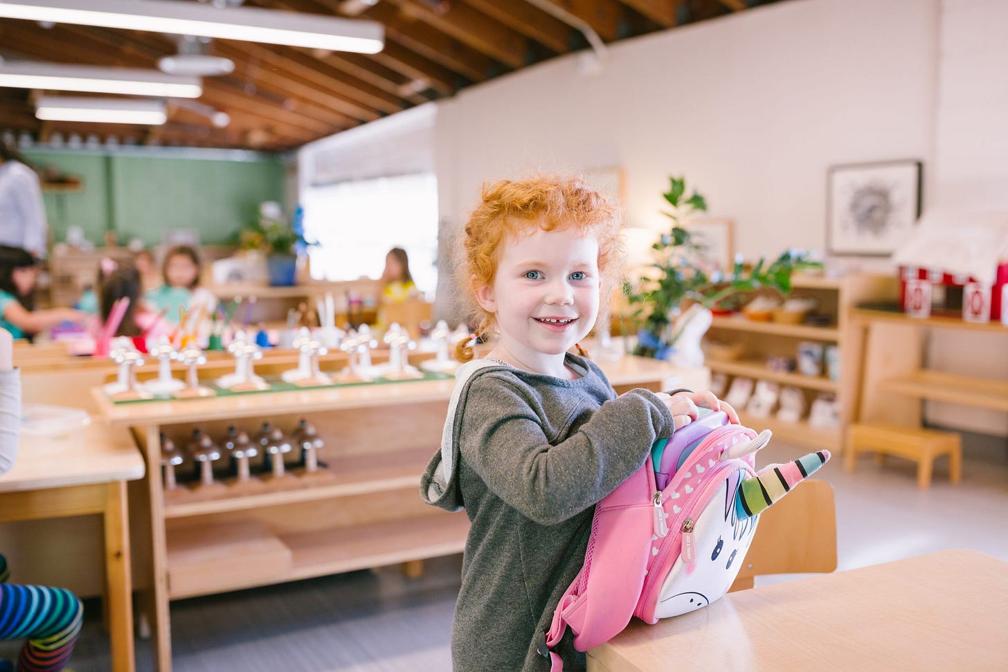 Young girl with curly red hair smiling in a Montessori classroom, holding a pink unicorn backpack