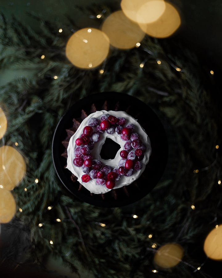 An overhead shot of vintage Bundt pants on a green background. A photo of orange carrots on a green background. A photo of shredded carrots in the foreground and the rest of the ingredients for Carrot and cranberry gingerbread cake. An overhead shot of the finished bundt cake with cream cheese frosting and sugared cranberries on top with white twinkle lights in the foreground and background. Another photo of the finished carrot and cranberry gingerbread cake surrounded by pine garland and white twinkle lights. 