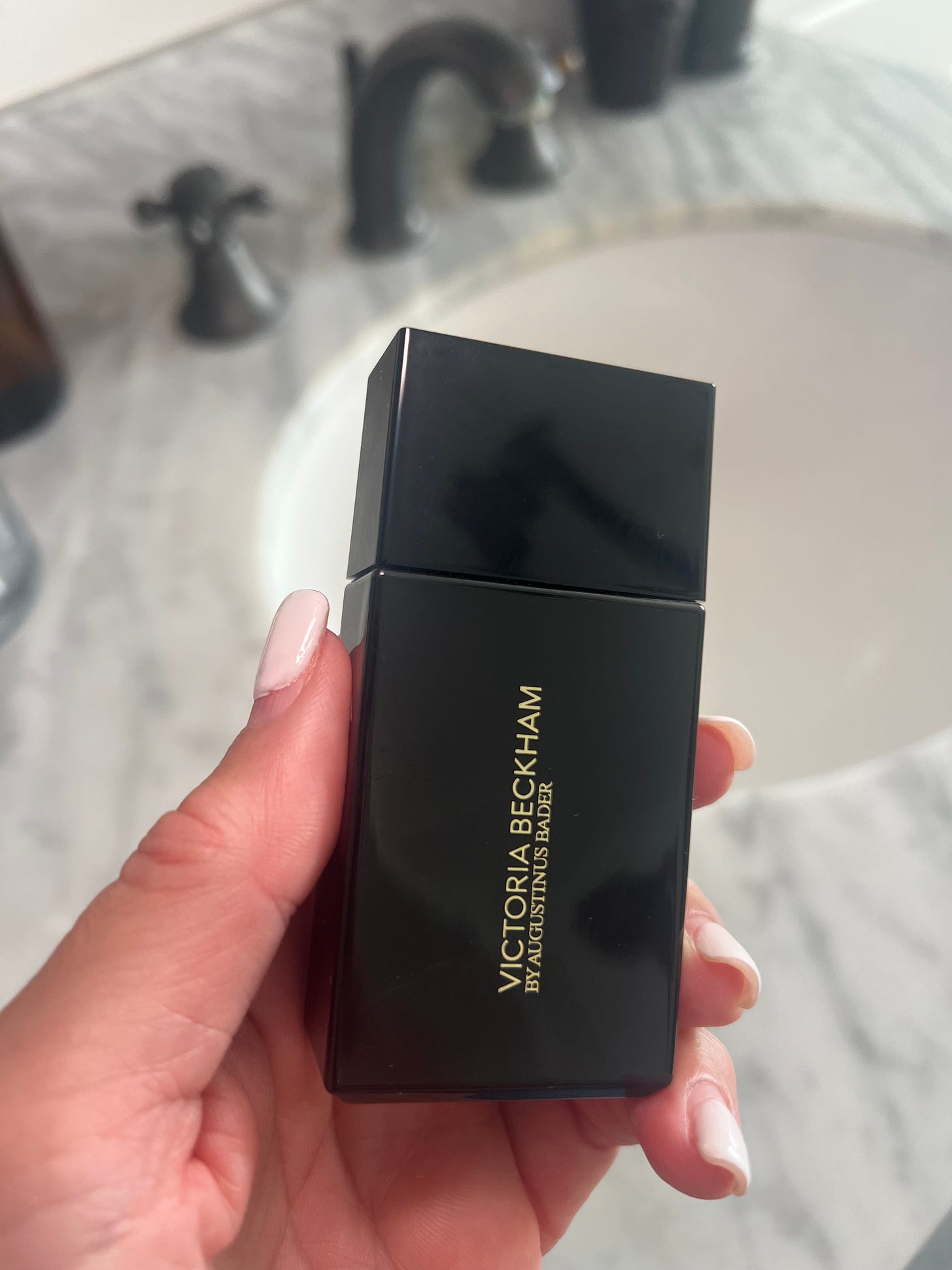 Close-up of Donna Francis holding the Victoria Beckham Foundation Drops bottle with its black and royal blue packaging against a bright, white marble sink.