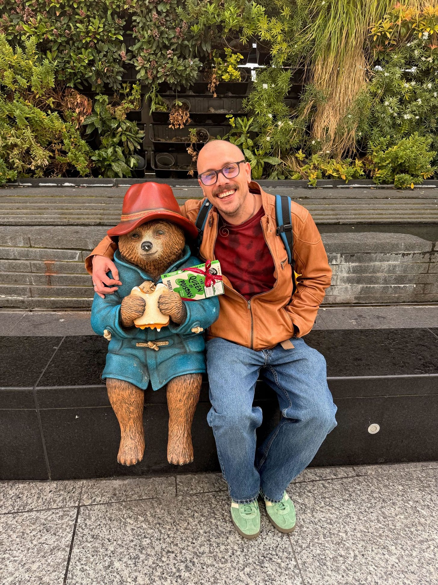 A photograph of Kris sitting on a bench with his arm around a Paddington Bear statue.