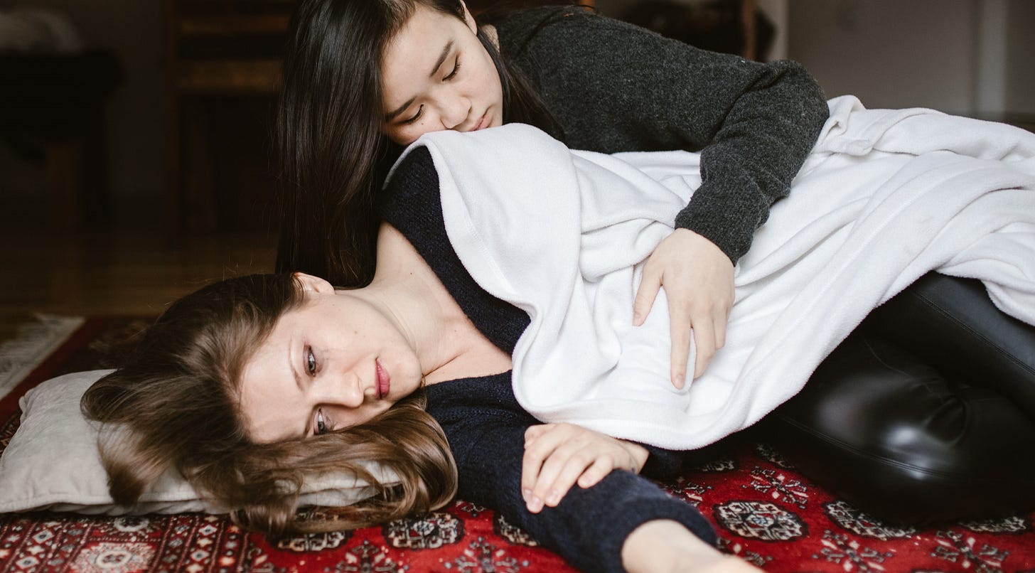 One woman holds another, who is despondant, as she lies on the floor.