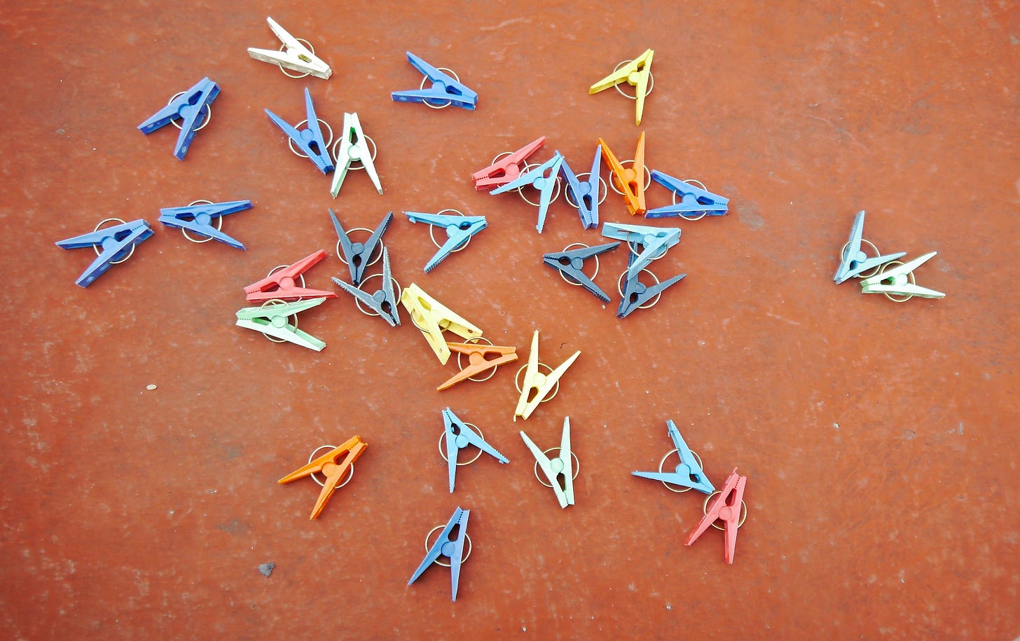 Una visione dall'alto di un assortimento di mollette colorate sparse su una superficie marrone rossiccia. Le mollette sono di vari colori, tra cui blu, giallo, bianco, arancione, rosso, verde chiaro e grigio scuro.