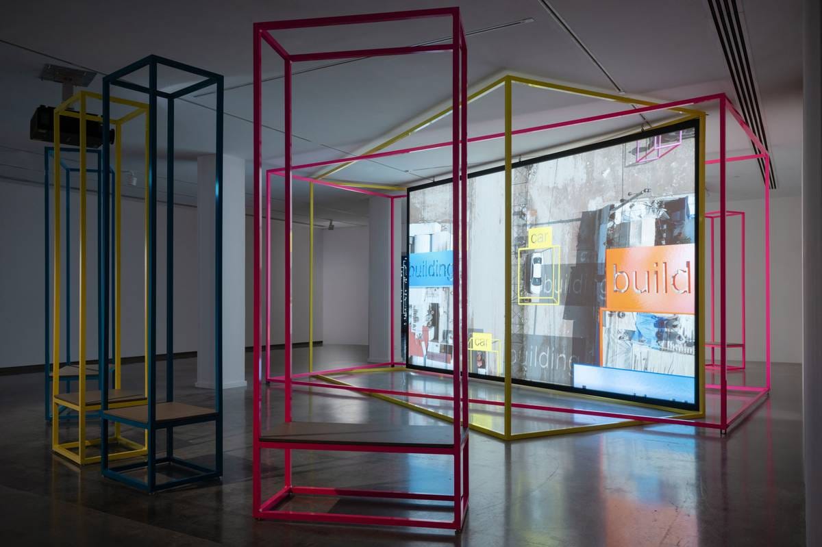 An installation image of a screen playing a video with painted metal structures in front of it. An installation image of a screen playing a video with painted metal structures in front of it.
