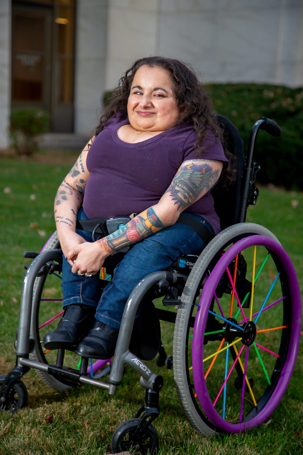 A full portrait of Valarie where she sits in her wheelchair outside of the Detroit Public Library. She has medium length brown curly hair parted in the center. She wears sparkly, pinkish eyeshadow and a rosy lip color. Her t-shirt is purple with a scoop neck. She is wearing jeans with black boots. Her tattooed arms are in front of her with her hands resting in her lap. Her wheelchair is manual, with purple push rims and rainbow spoke covers on the wheels.