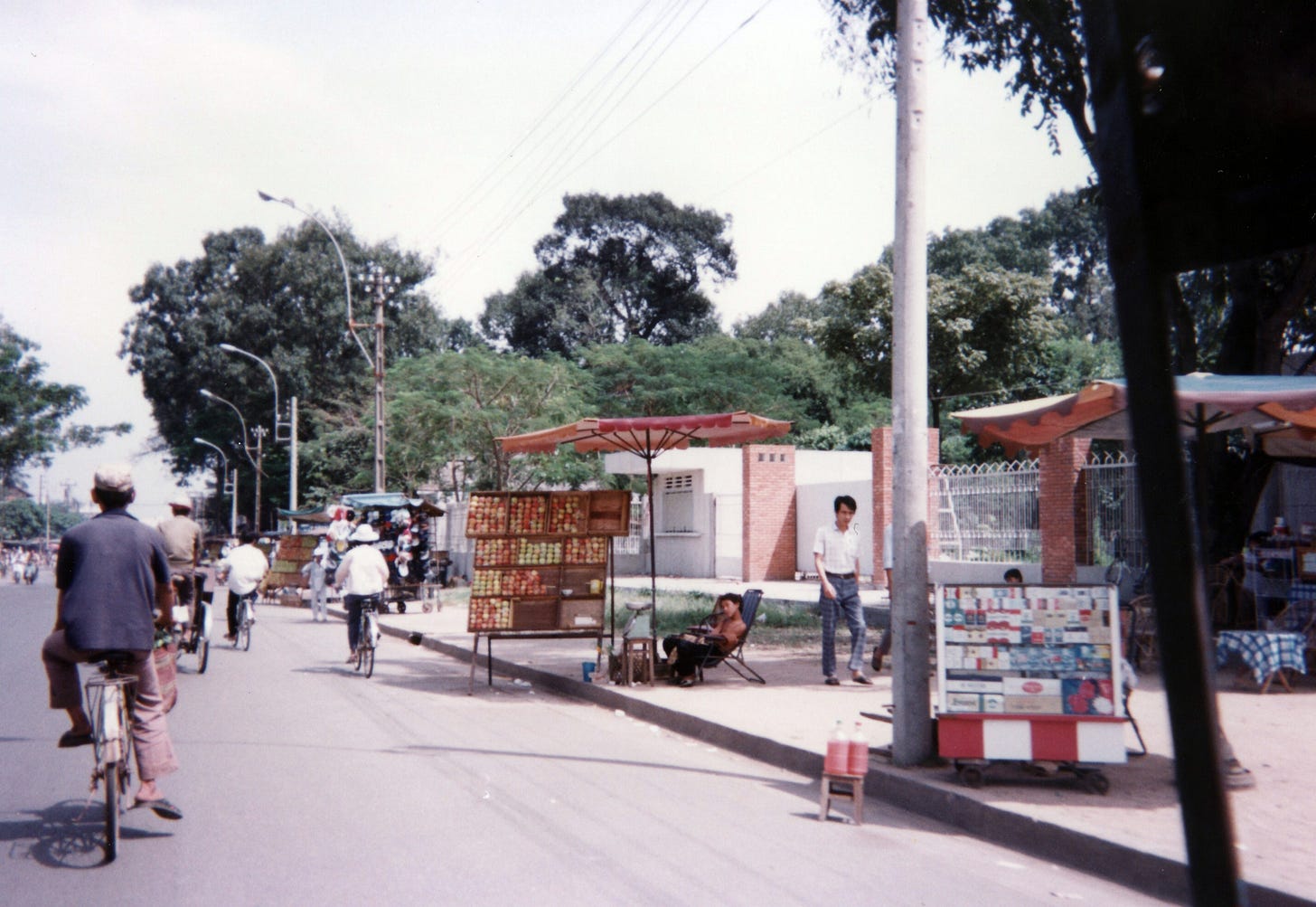 A street scene in Vietnam more than 30 years ago. People are selling goods from stands at the side of the street and folks are riding bicycles. The photo is grey and faded. 