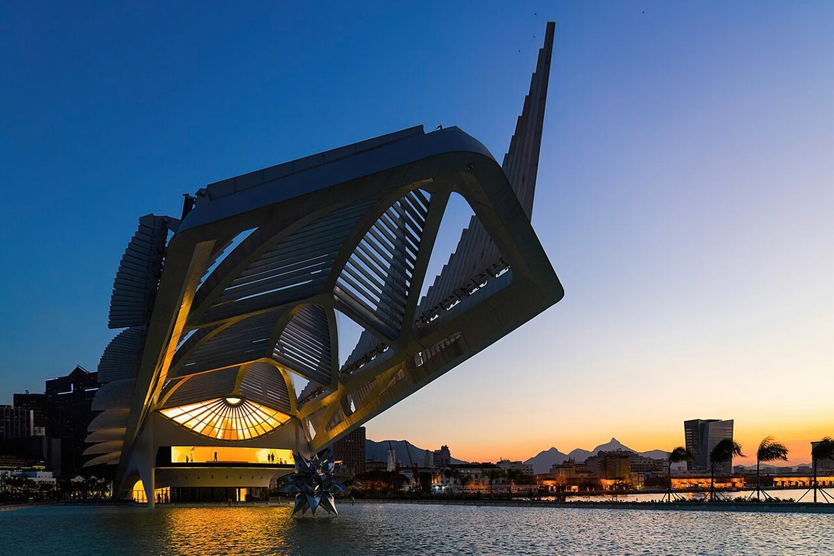 A futuristic white architectural structure resembling a stylized spaceship or skeleton sits at the edge of a body of water at sunset. The building has sharp angles and a large overhanging canopy with slatted panels. The warm glow of the setting sun illuminates the horizon, casting reflections of the building and the sunset colors on the water's surface in the foreground. In the distant background, silhouettes of palm trees, city buildings, and mountains are visible against a blue and orange sky.