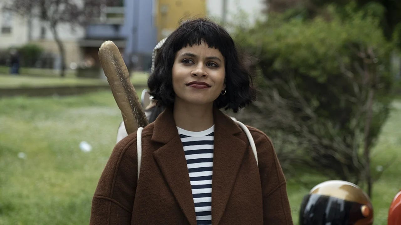 Cena de Atlanta, com a personagem Van. Uma. mulher negra de cabelo chanel e franja, blusa listrada e uma mochila nas costas de onde sai a ponta de uma baguete Cena de Atlanta, com a personagem Van. Uma. mulher negra de cabelo chanel e franja, blusa listrada e uma mochila nas costas de onde sai a ponta de uma baguete