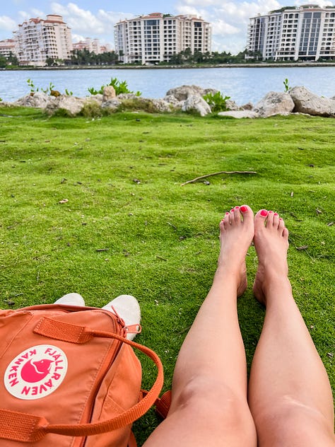 Photo of a hand holding up an illustrated book entitled 'Trees,' in a living room with a red bookcase and a tv in the background; mirror selfie photo of woman in a short dress and cap and carrying a bag; photo of a tree with several orchids of different colors along a sidewalk in front of a building; photo of a coastline with water in the background and a city skyline in the distant background, with a towering art sculpture and palm trees in the foreground; a photo of a woman's legs and painted toes sitting on green grass with a backpack on her left and a rocky shore and water from a bay in front of her; photo of a white stone archway with ivory and patterned tiles inset, and palm leaves on the ground below, with patches of light and shadow against the wall; a convertible car with paintings and the words 'Everybody LOVE everybody' and bumper stickers that say things like 'Life Without Art is Stupid'; a photo of a head-on view of a baby blue with yellow trim lifeguard station on the sand with the ocean behind it; an overhead photo of a stack of magazines, scissors, and magazine clippings for a collage, on top of a clear glass coffee table and tile floor and rugs.