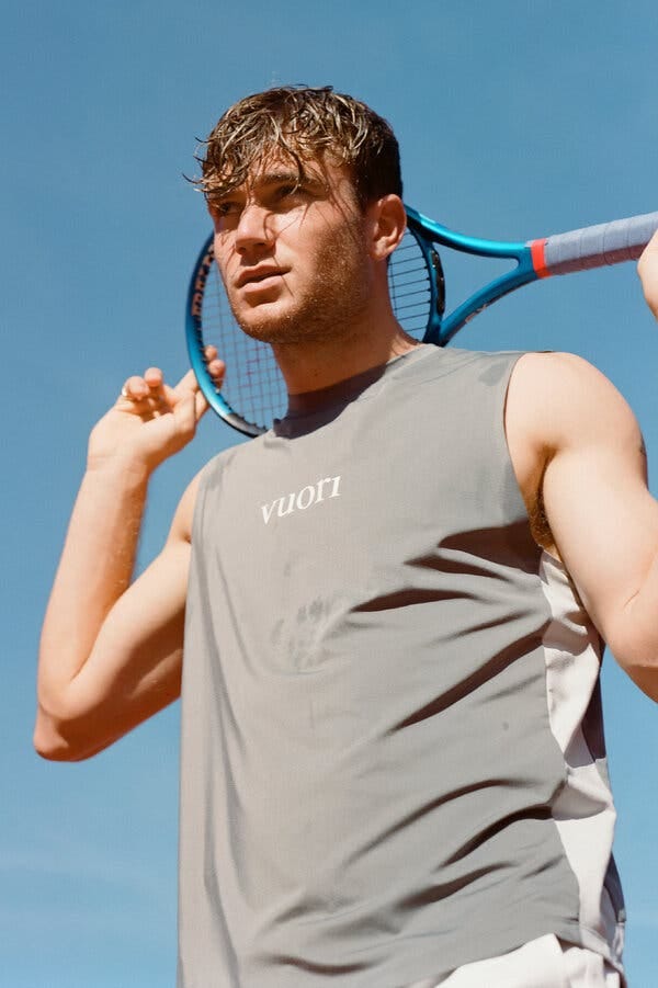 A man in a sleeveless gray shirt that says Vuori on it holds a tennis racket behind his head.