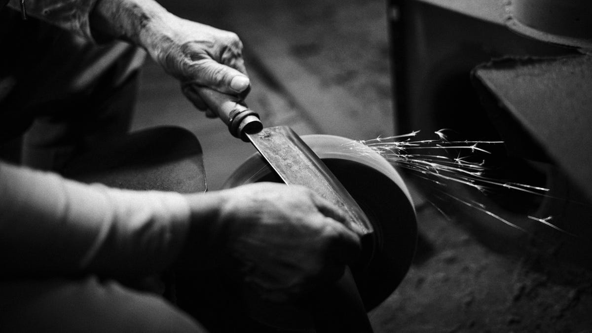 Hands sharpening a blade on a grinding wheel, with sparks flying, in a black and white image showcasing a traditional crafting process.