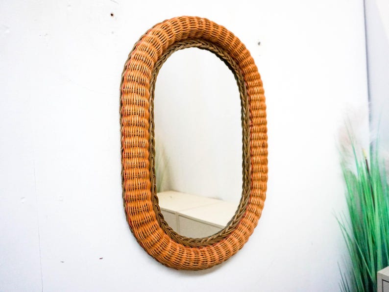 May include: An oval-shaped wicker mirror with a natural brown wicker frame and a light orange wicker border. The mirror is reflecting a white surface.