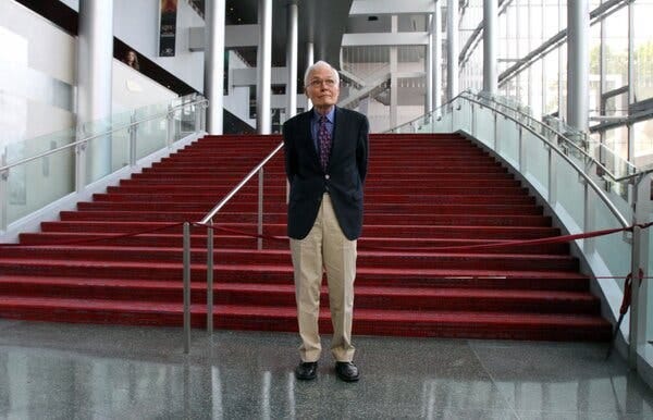 Mr. Jenkins stands with his hands behind his back, wearing a dark blazer and khaki pants, in front of a wide red staircase.